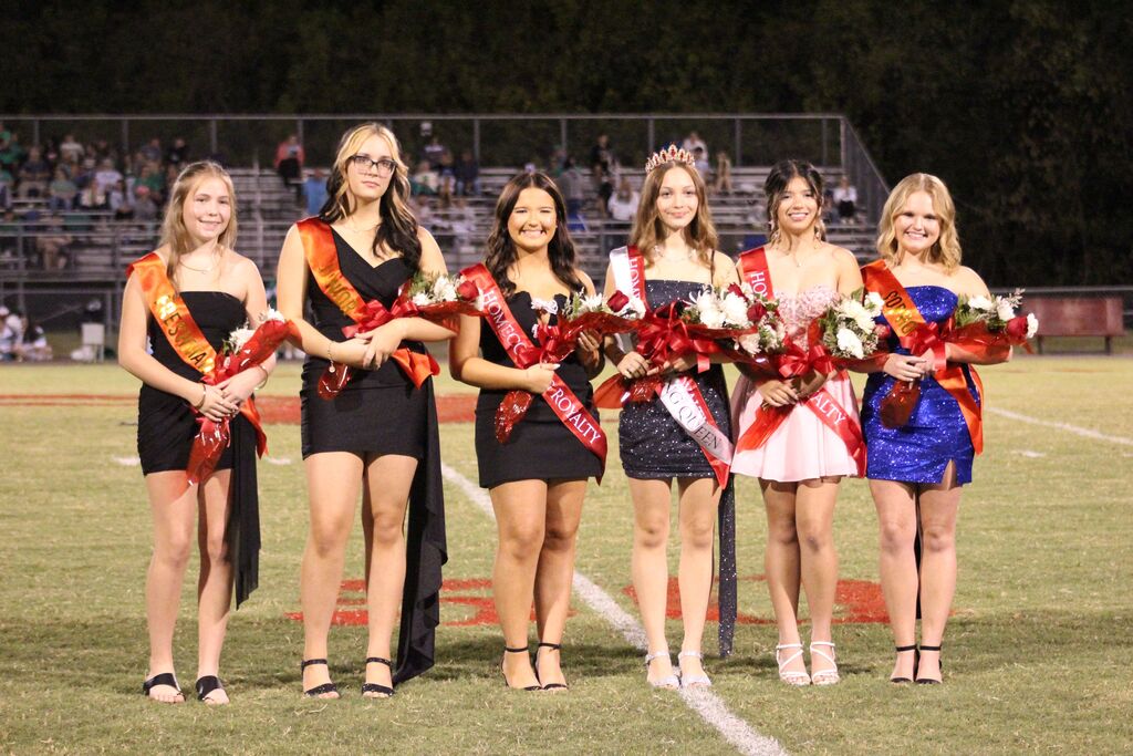 Six homecoming court members stand on a football field during a nighttime ceremony, wearing formal dresses, sashes, and holding bouquets of flowers. One court member wears a crown, while stadium bleachers and spectators are visible in the background.