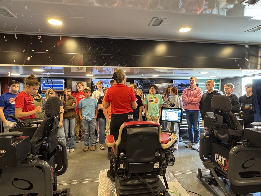 A group of OCMS students stands among heavy equipment simulators inside the Be Pro Be Proud mobile workshop as the adult leader addresses them.