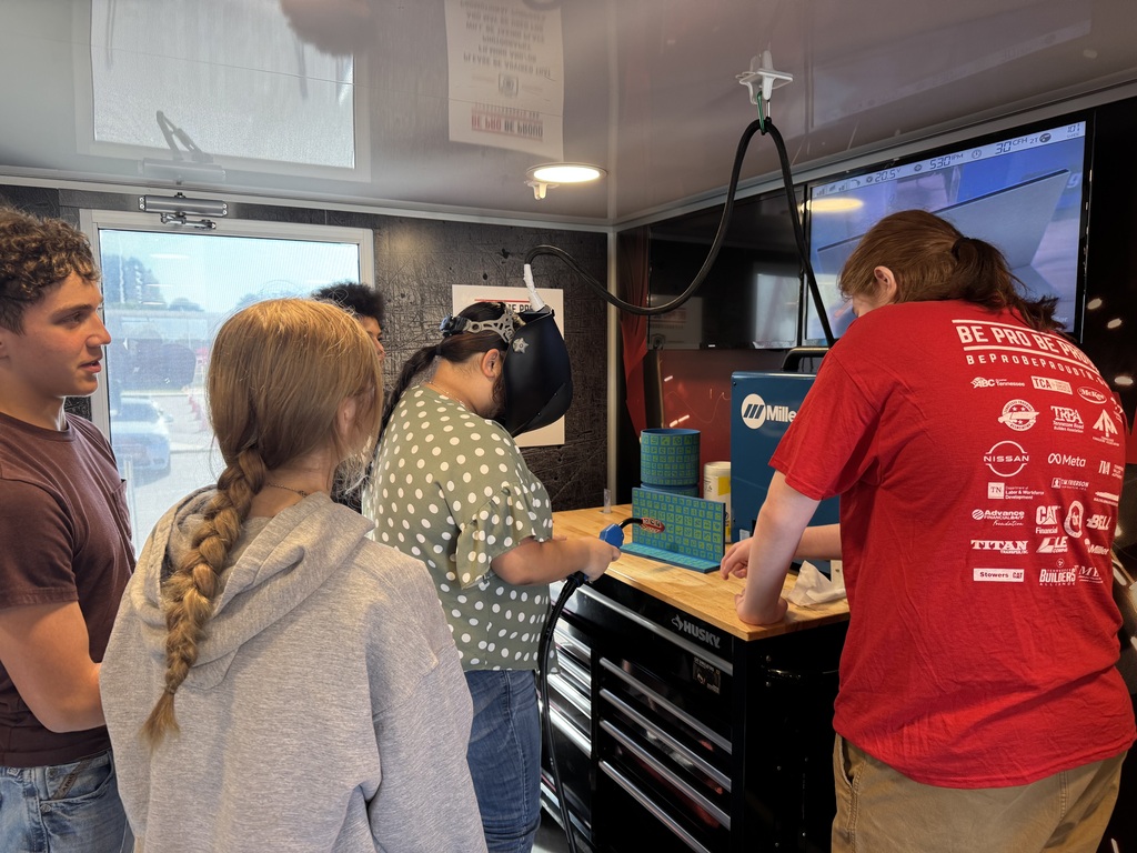 A student wearing a virtual reality headset practices at a workstation while two classmates watch and a student ambassador assists.