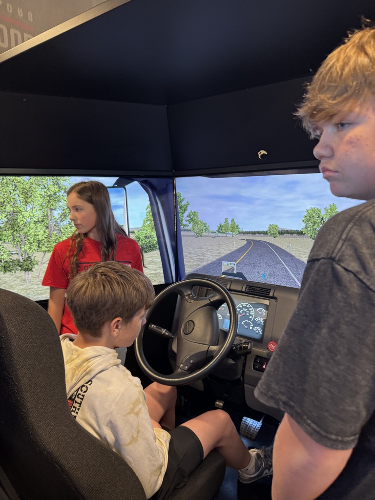 A student sits at the steering wheel of a truck-driving simulator, facing screens displaying a roadway. A student ambassador and another classmate stand nearby.