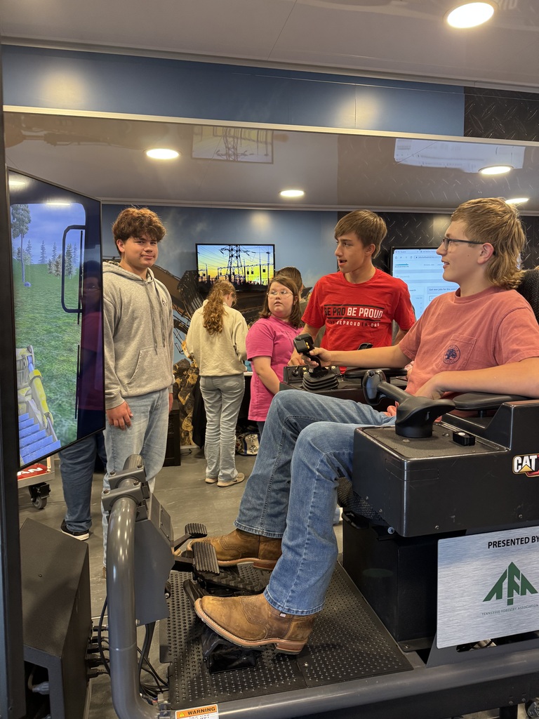 A student operates the joysticks of a CAT heavy equipment simulator while a student ambassador and classmates watch the screen.
