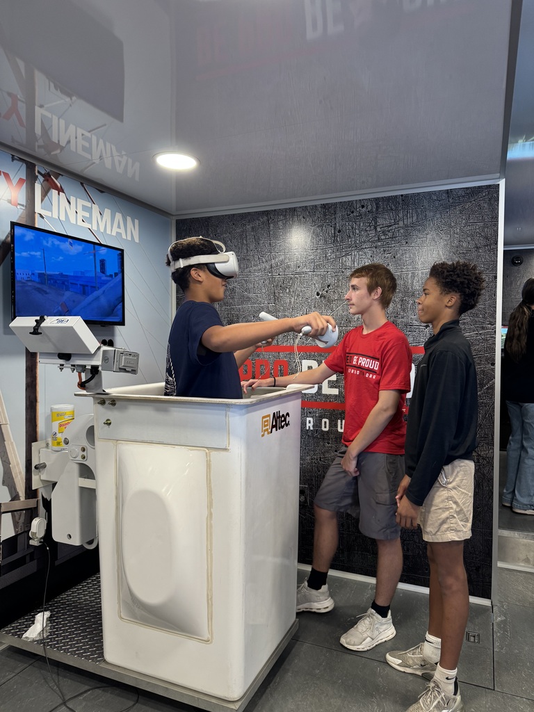 A student wearing a virtual reality headset reaches forward at the lineworker simulator while a student ambassador and another classmate stand nearby.