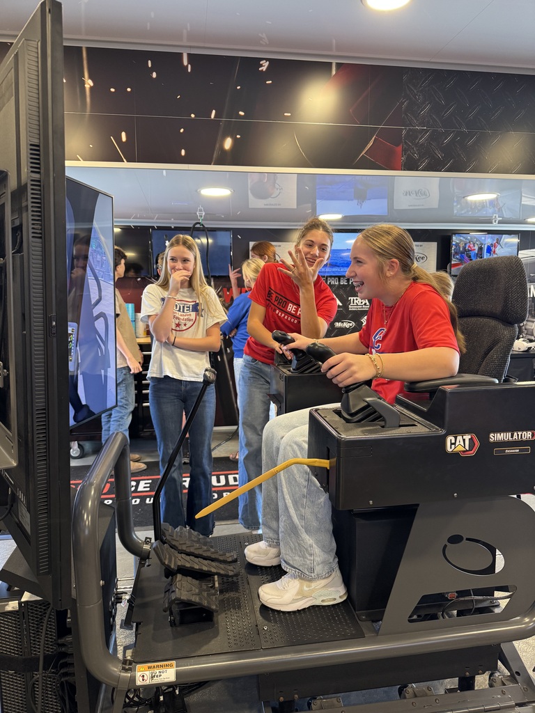A student seated in a CAT heavy equipment simulator smiles while using the controls. Two students stand nearby inside the Be Pro Be Proud mobile workshop.