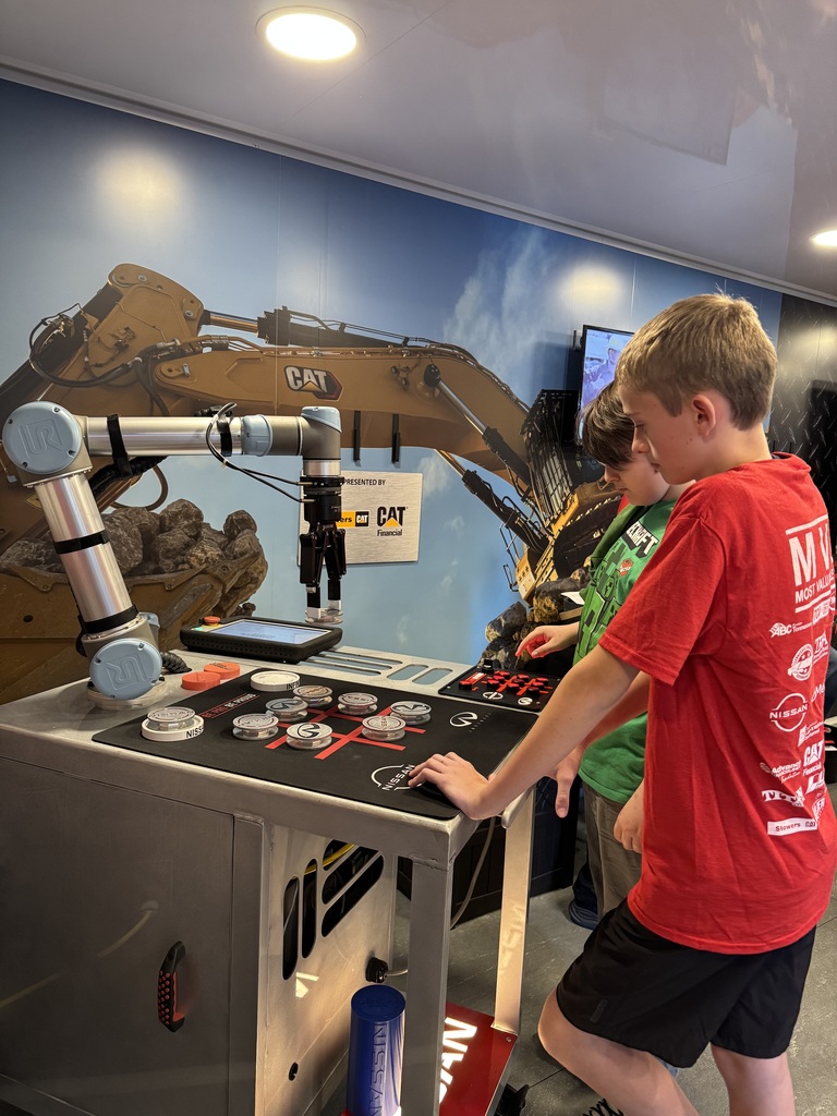 A student explores a control panel beside a robotic arm while a student ambassador watches. An excavator illustration covers the wall behind them.