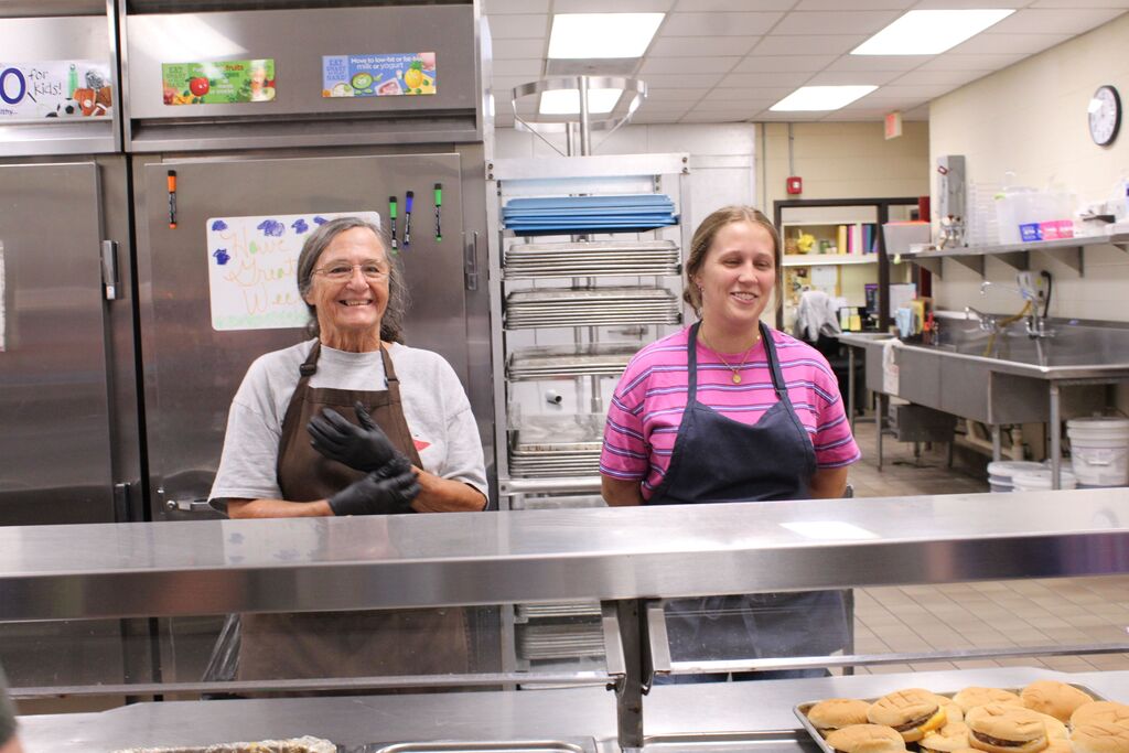Two smiling cafeteria employees oversee students filling their lunch plates.