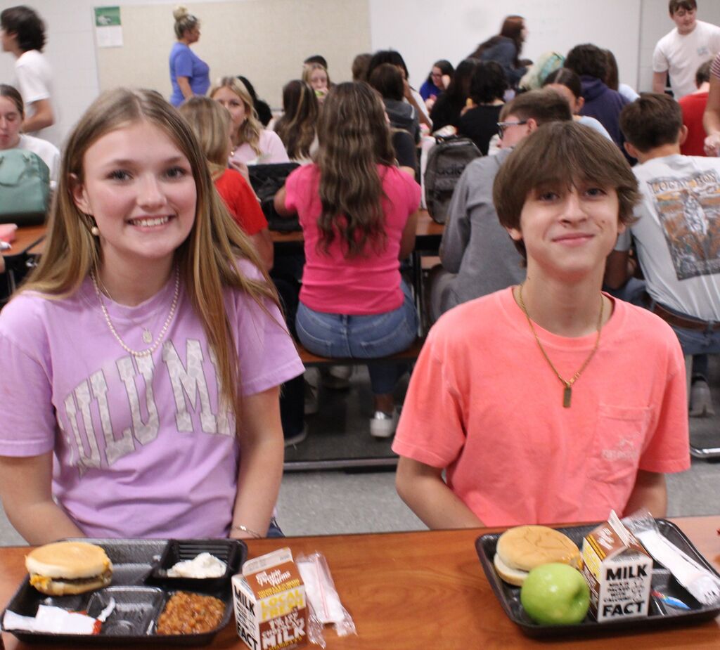 A boy student and girl student sit at a cafeteria table enjoying their school lunches.