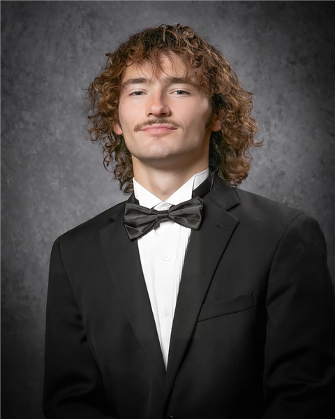 Boy student in a black tuxedo smiles at the camera.