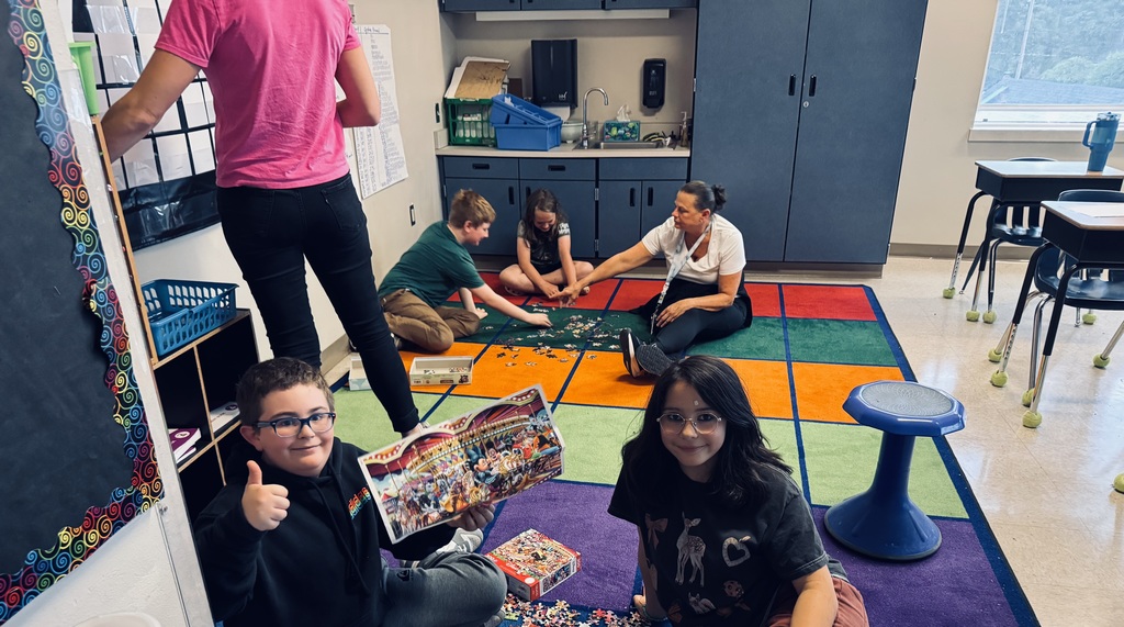 Four students and one teachers sitting in two groups on the floor putting two puzzles together in a classroom.  A teacher is standing in the frame fixing a  board
