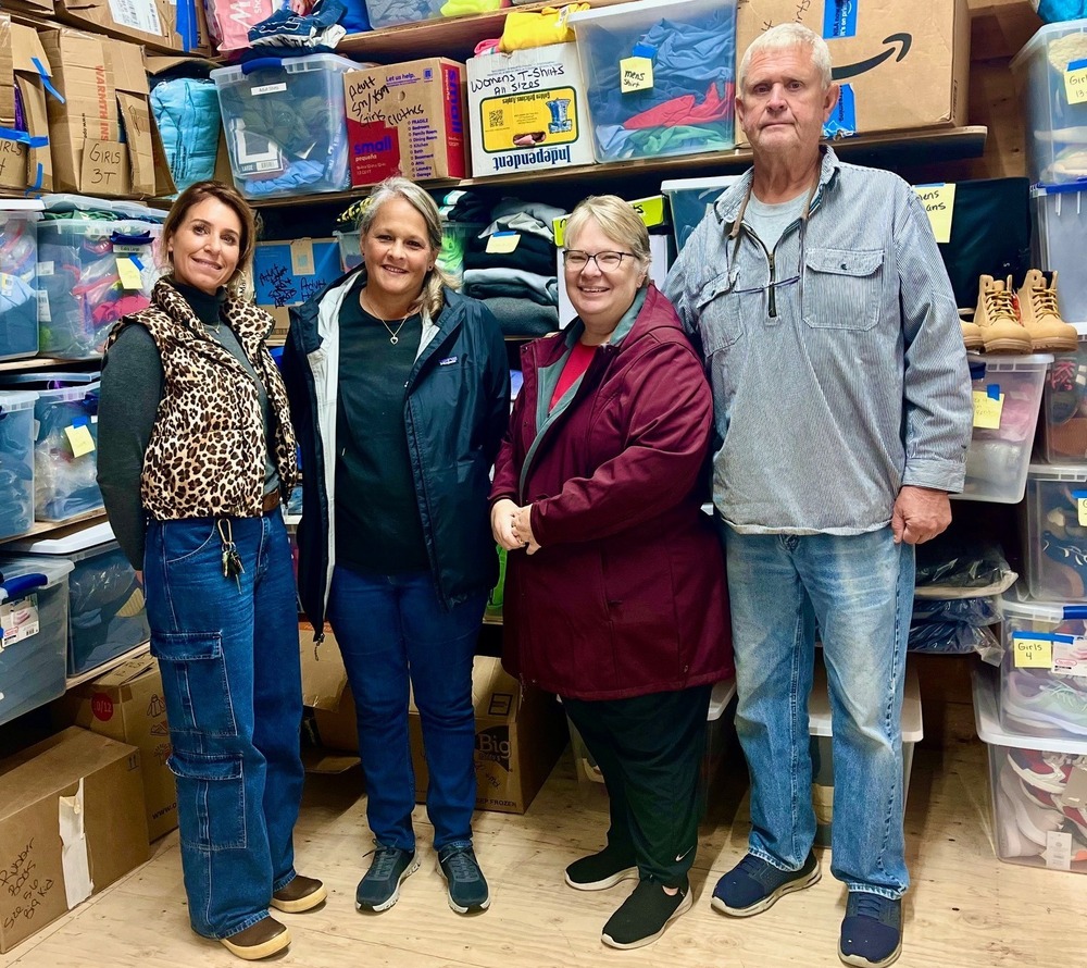 4 adults standing in a room with boxes and bins of clothing behind them.