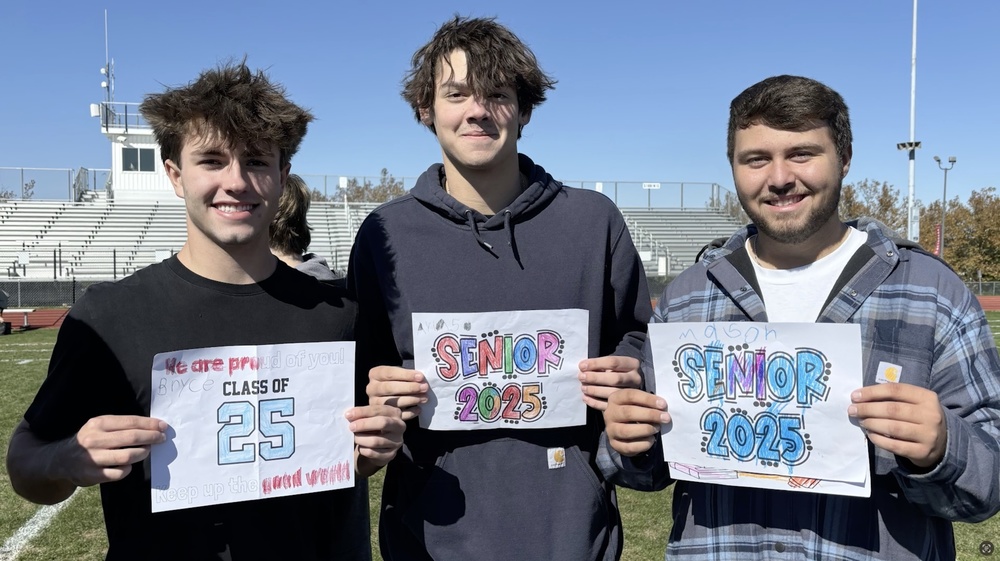 three male students outside on the football field, holding up signs about being seniors
