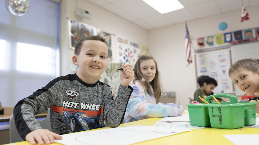 little boy holding marker and smiling at camera, sitting at yellow table coloring with two classmates