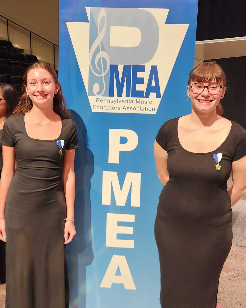 Two students dressed in black concert attire stand smiling beside a blue PMEA (Pennsylvania Music Educators Association) banner inside a performance hall. They are wearing participation medals, with auditorium seating visible in the background.