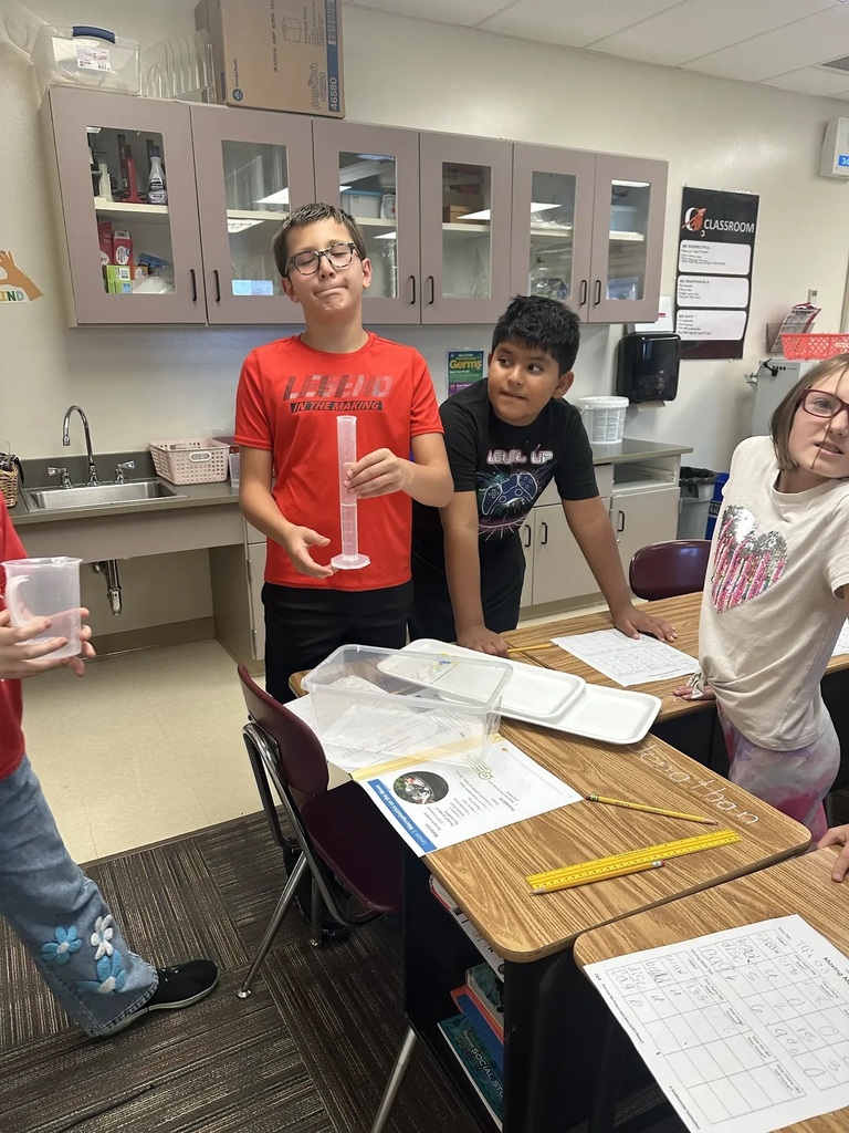 student holding plastic beaker in front of desk with other students looking on