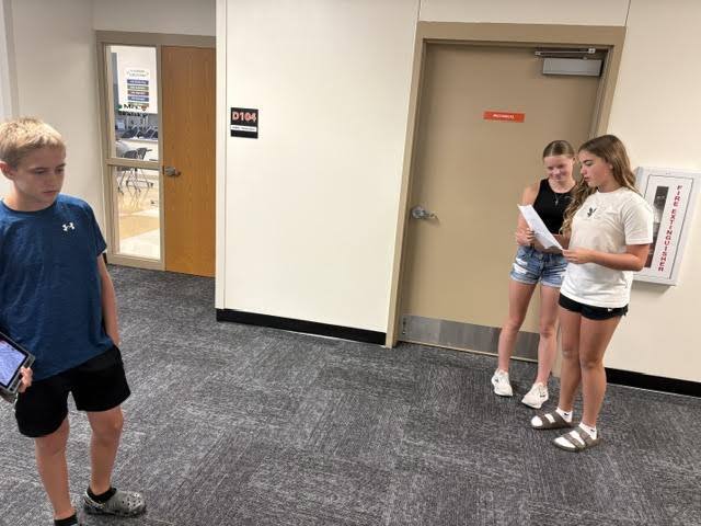 students standing in hallway looking at paper, while boy holds ipad