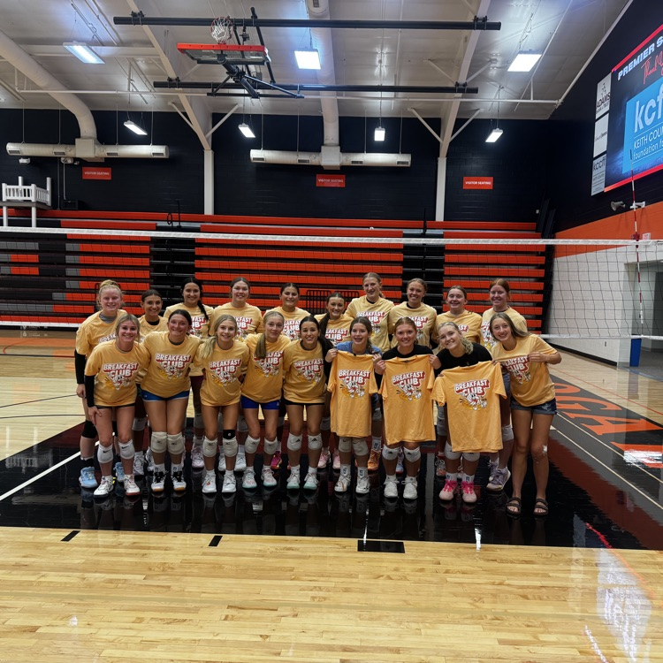 two rows of volleyball players holding a yellow breakfast club T-shirt, standing in front of the volleyball net