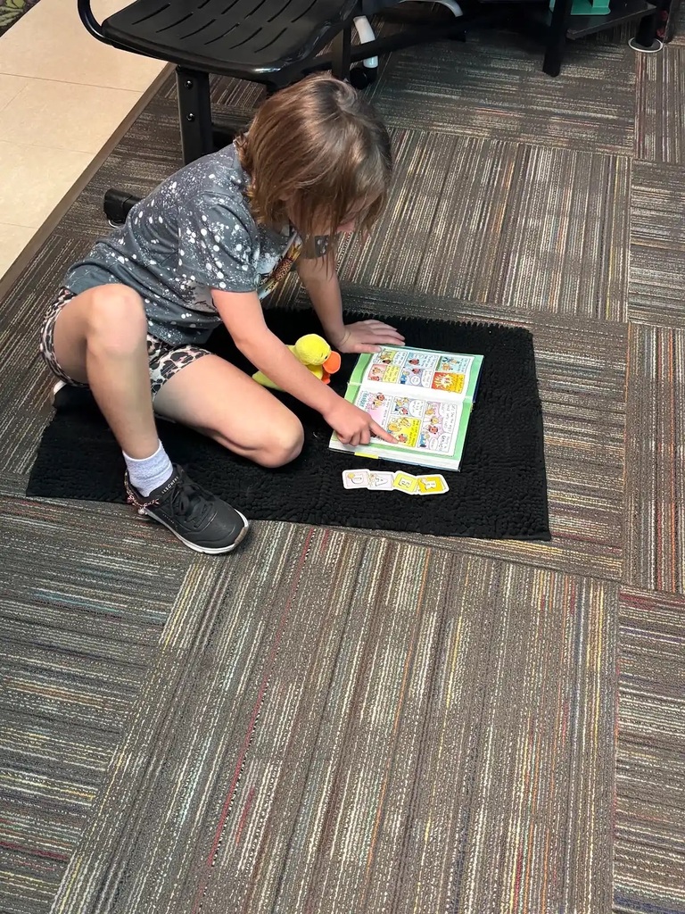 student sitting on a rug while pointing to a page in a book