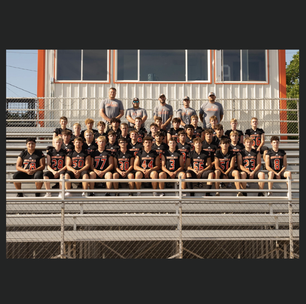 football team sitting on bleachers wearing black jerseys with coaches standing in theback 