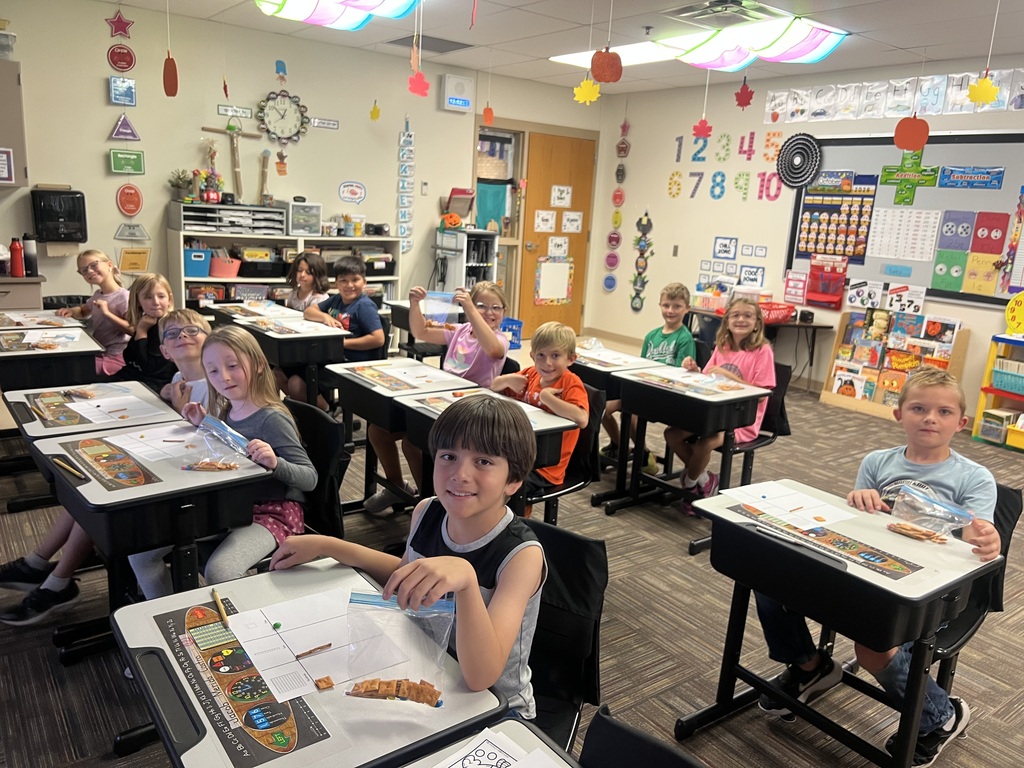 students sitting in desks in a classroom holding a bag of snacks smiling at the camera