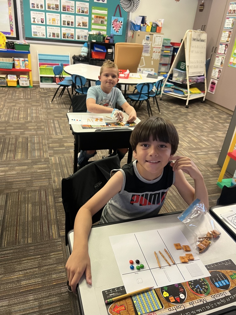 students sitting in desks in a classroom holding a bag of snacks smiling at the camera