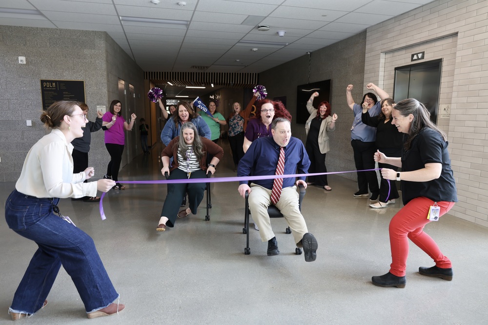 People racing down the hallway on desk chairs, and others cheering them on. As well as a finish line being held. 