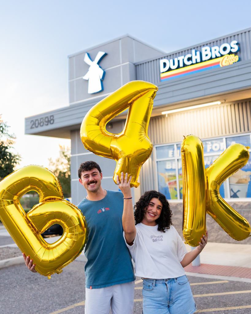 Two individuals holding gold balloons in front of Dutch Bros store