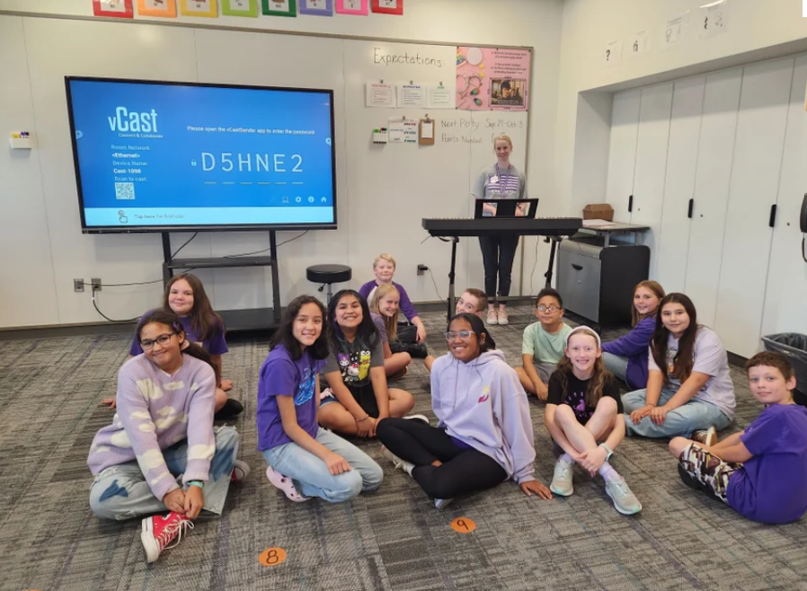Students sitting on carpet, music teacher with new piano behind
