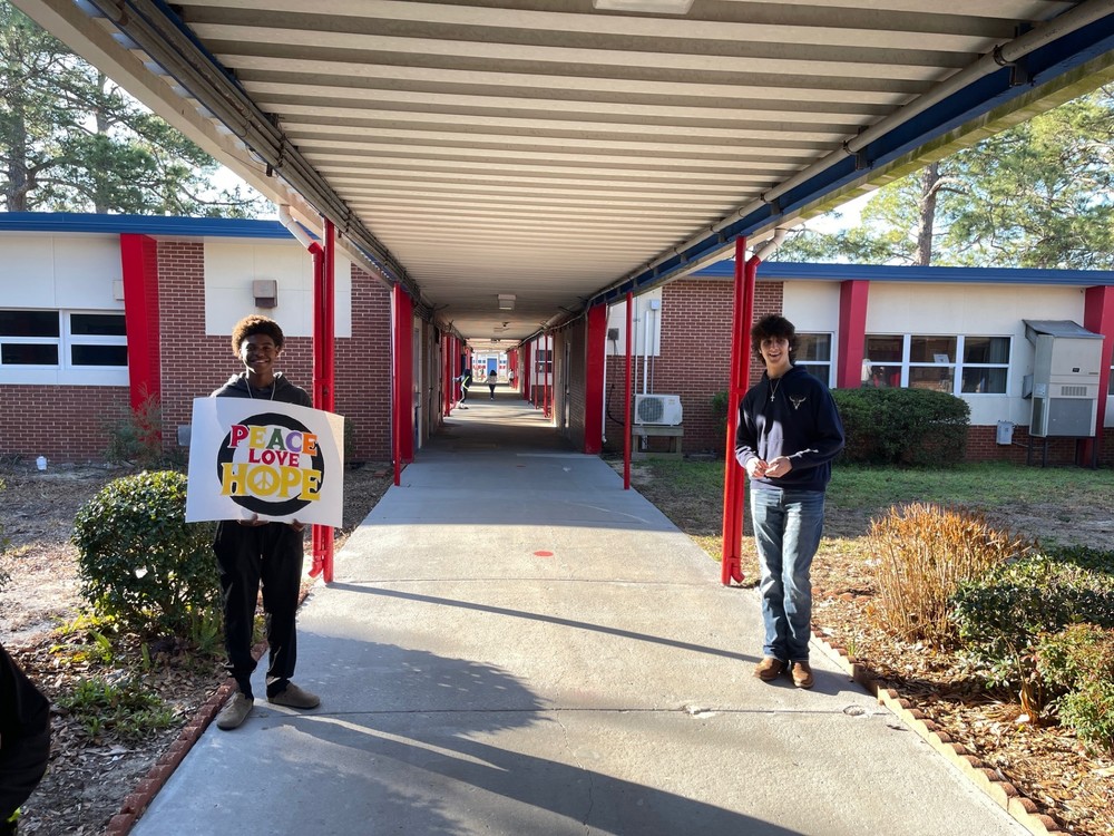 A covered outdoor walkway at a school, lined with red support poles and bordered by brick buildings. Two people stand on either side of the walkway entrance. One holds a colorful sign that reads “PEACE LOVE HOPE.” Sunlight casts long shadows across the concrete path.