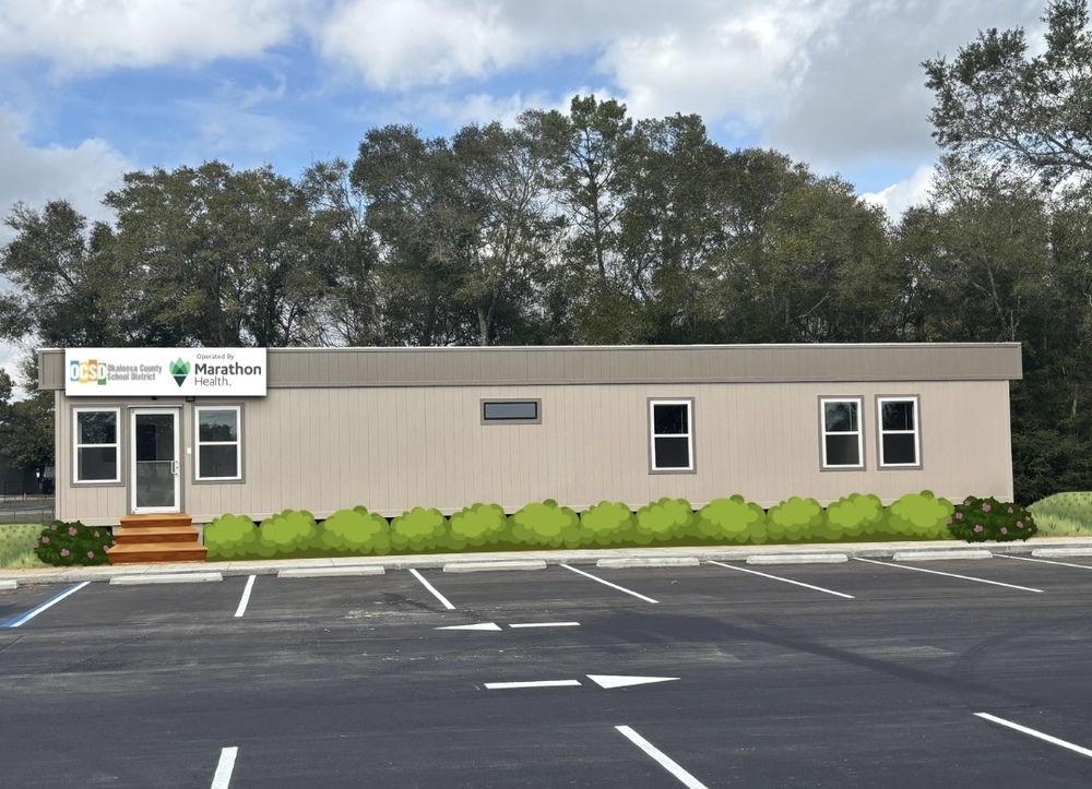 A beige modular building with several windows and a small stairway sits in front of trees. A sign on the left displays the Okaloosa County School District and Marathon Health logos. Green shrubs line the front, and an empty parking lot is in the foreground.