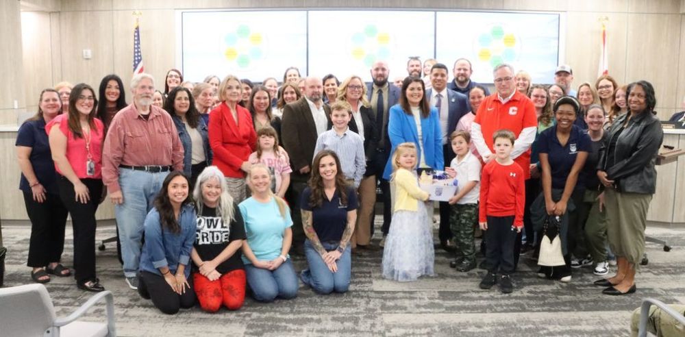 Brittany Young, named Principal of Walker Elementary School, is pictured with a large group of adults and children at the School Board meeting. They are standing and kneeling in rows on a patterned carpet, with a large screen displaying colorful graphics behind them and U.S. flags on either side. One child in a light blue dress stands near the center holding a box, while others are dressed in casual or school attire.