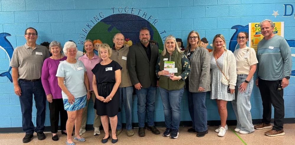 Shalimar Library Board of Directors, Mayor Mark Franks, Principal Amy Klugh and Shalimar Elementary School Administration stands together in a single row inside a school hallway. They are posing for a group photo in front of a blue mural featuring a globe and the words “Working Together.” One person near the center holds a booklet or certificate. The group is casually dressed, and a colorful school poster is visible on the wall to the right.