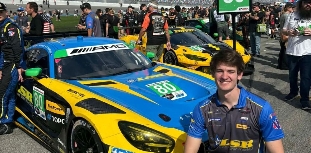 A race car painted blue and yellow with the number 80 displayed on the hood is parked on a racetrack grid surrounded by a crowd. Another race car painted yellow and black is positioned behind it. People wearing racing team uniforms and event apparel stand and walk around the cars. Jackson Shirey, in a team shirt, is positioned in the foreground near the front of the blue-and-yellow car. A large crowd fills the background along the track.