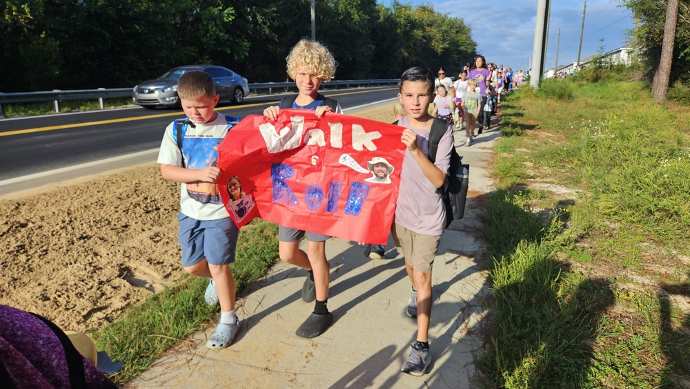 A large group of Riverside Elementary students, families, and staff walk together along a sunny sidewalk for National Walk to School Day. Three students lead the group holding a bright red banner that says “Walk & Roll,” decorated with photos and glitter letters. Cars are visible on the road beside them, and more walkers can be seen stretching into the distance behind them.