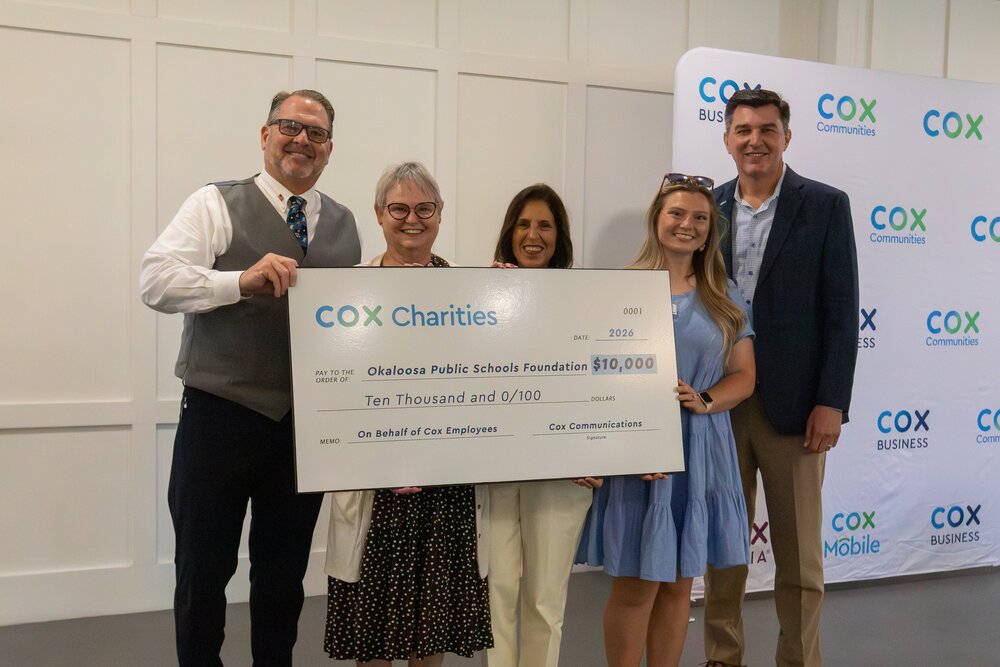 Five individuals stand indoors in front of a Cox Communications backdrop, holding a large ceremonial check from Cox Charities made out to the Okaloosa Public Schools Foundation for $10,000 (dated 2026). The group is positioned in a well-lit room with a light-colored paneled wall, and the check prominently displays “Cox Charities,” “Okaloosa Public Schools Foundation,” and “Ten Thousand and 0/100 Dollars.”
