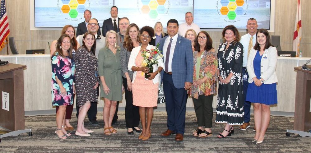Mrs. Ross stands in the middle of a group of 15 people, holding a bouquet of flowers, in the School Board meeting room in front of a large presentation screen. The group is positioned in a semi-circle on a carpeted floor with desks, microphones, and an American flag visible behind them. Two large display screens in the background show colorful hexagon graphics.