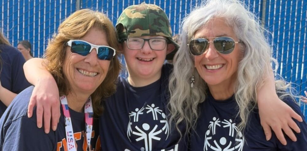 Three people in Special Olympics shirts pose together outdoors in front of a blue fence.
