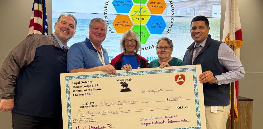 Pictured above left to right: Assistant Superintendent John Spolski, Director Stephen Anderson, Pamela Woodward and Nanette Roaden of the Fort Walton Beach Moose Lodge, along with Superintendent Marcus Chambers stand in the Board Room holding an oversized ceremonial check. The check is made out to Okaloosa County Schools for $6,002 and is presented by the Loyal Order of Moose Lodge 2193 and Women of the Moose Chapter 2320. Behind the group is a large colorful district emblem on the wall and an American flag and a Florida state flag on either side.