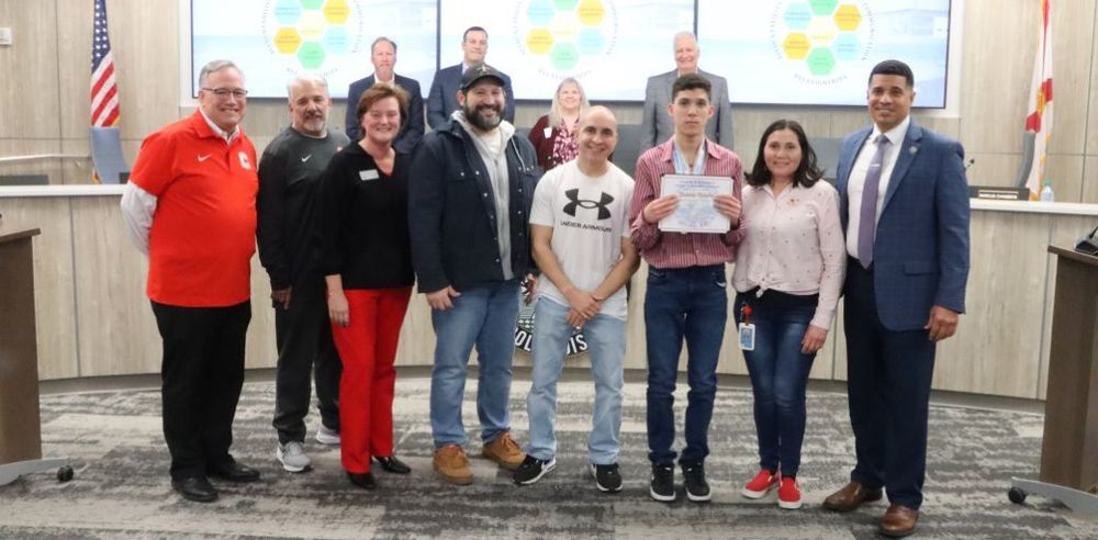 Donovan Morales, Crestview High School, Wins FHSAA Class 3A Boys Swimming 50-meter and 100-meter Freestyle State Championships. He is pictured holding a plaque with a large group who stands together at the School Board meeting. A color display screen and flags are visible behind them.