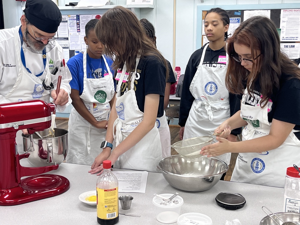 Students wearing aprons participate in a hands-on culinary activity in a classroom kitchen, working together to prepare ingredients. One student uses a red stand mixer while another sifts flour into a metal bowl, as an instructor guides them. Measuring tools, ingredients, and a recipe sheet are spread across the table, and posters and notices are visible on the wall in the background.