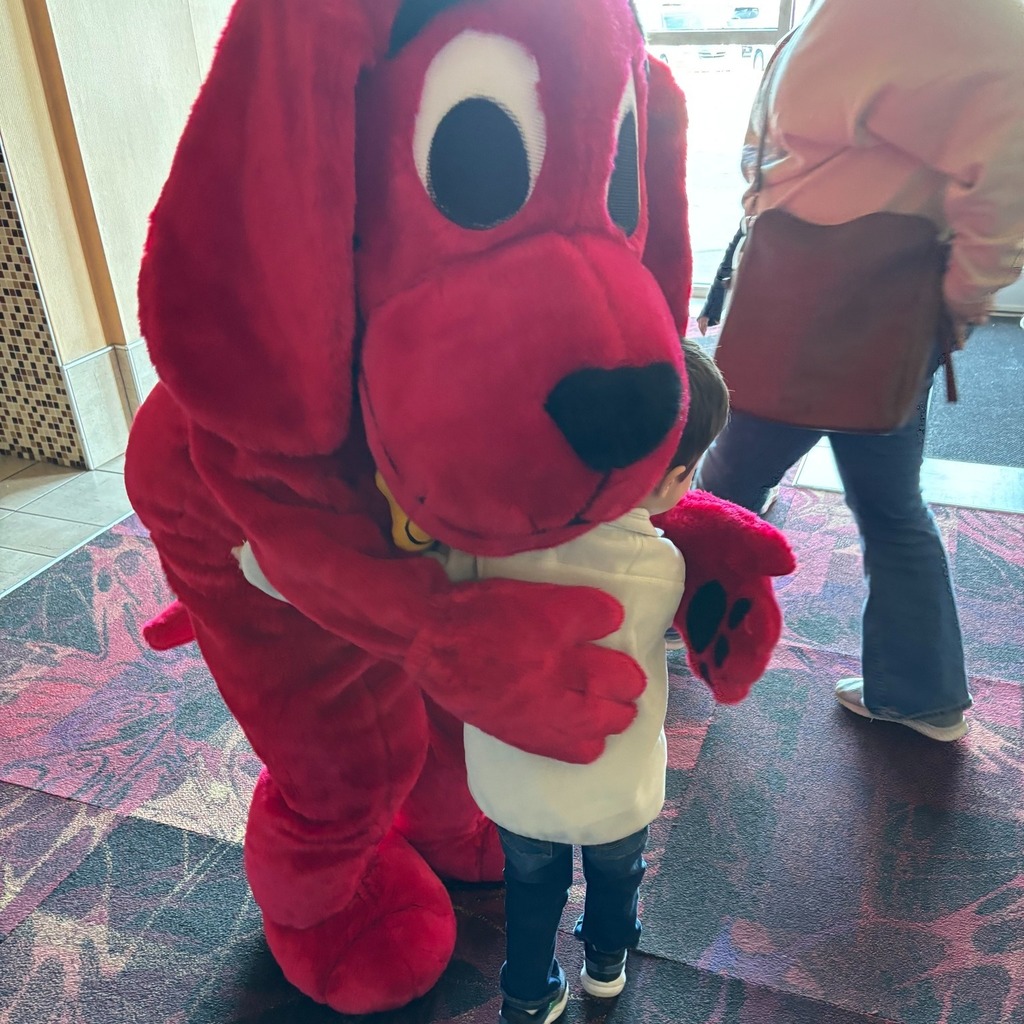 A person in a large red dog mascot costume hugs a young child indoors, while another adult walks nearby in a public lobby or hallway with patterned carpet and tiled walls.