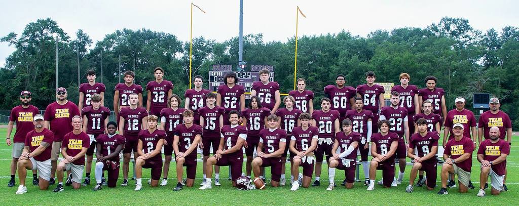 A football team in maroon “Gators” uniforms poses on a football field with coaches, arranged in rows, with goalposts and trees in the background during a team photo.