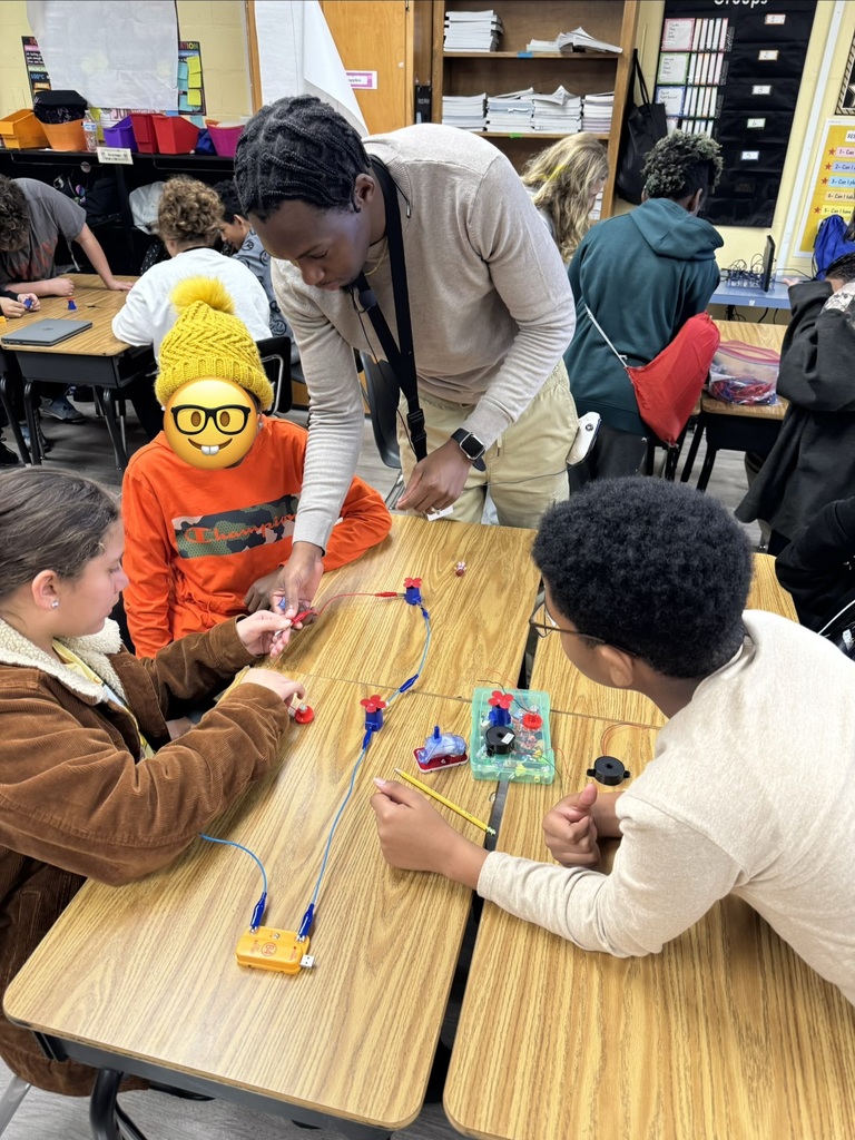 A classroom scene where Mr. Fraser is assisting a group of students working on an electronics or circuitry activity at a table. The students are gathered around components, wires, and small devices while other groups work at tables in the background. One student is wearing a bright yellow knit hat with a smiling face graphic overlaid.