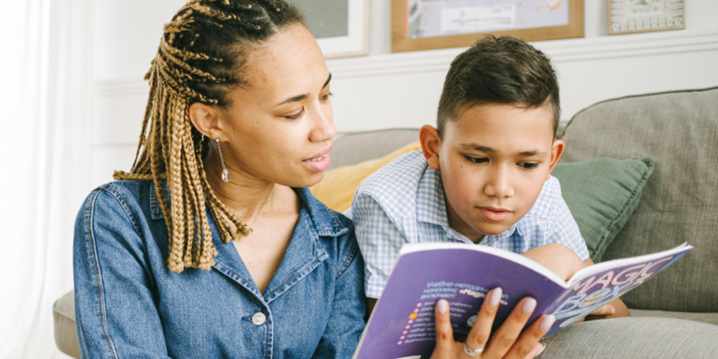 Two people sit together on a couch, looking at an open book held by the person on the left. The book has a purple cover with colorful text. A green cushion is visible behind them, and framed items hang on the wall in the background.