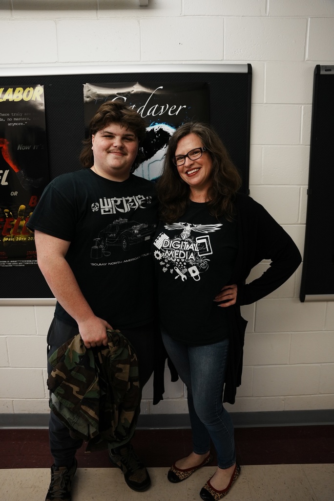 Own Atkinson pictured standing with his teacher in front of a bulletin board with several posters. Both are wearing black graphic T-shirts—one featuring a design with text resembling “LPRRE” and the other featuring a digital media–themed graphic. The background includes a white brick wall and dark floor tiles.