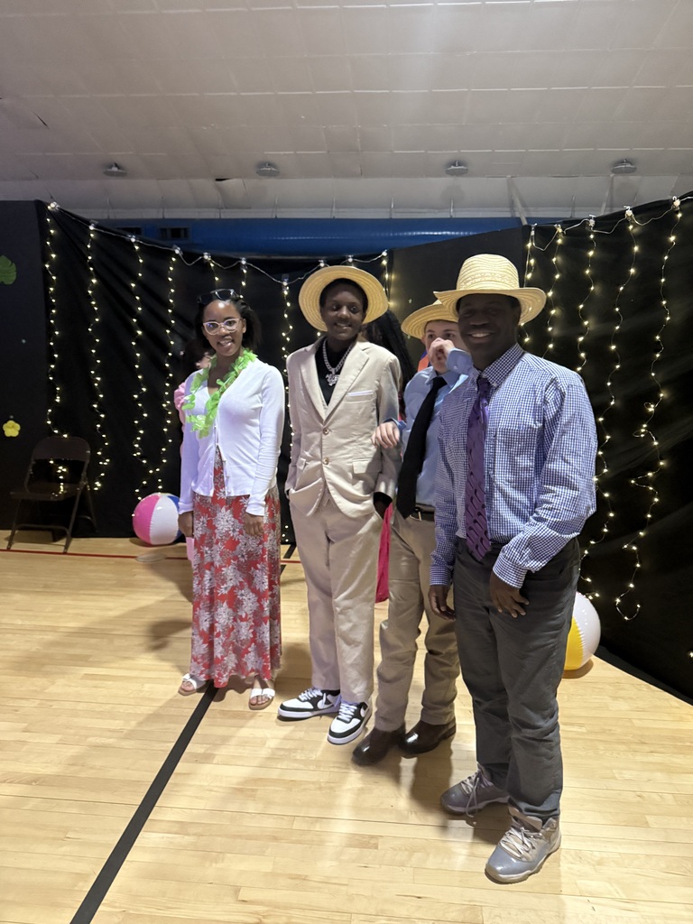 Four students stand together at prom wearing a mix of formal and tropical-themed outfits, including leis and straw hats. They are smiling and posing in front of a backdrop with string lights. Beach balls and decorations are visible on the floor, adding to the Hawaiian “Lilo and Stitch” theme.