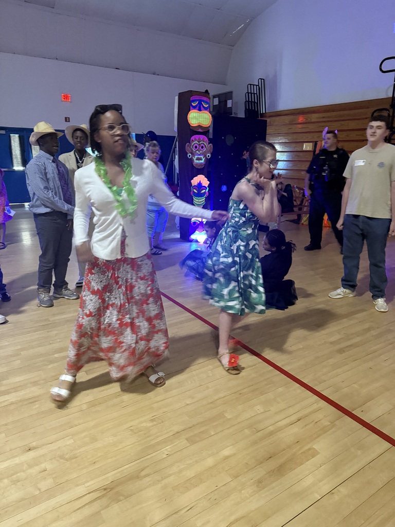 Students dance together on the gymnasium floor during a school prom, with decorations and other attendees visible in the background.