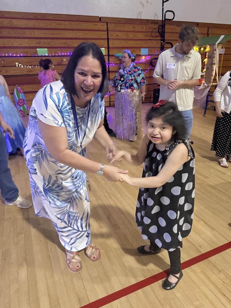A teacher dances with a student on the gymnasium floor during a school prom, with other students and decorations visible in the background.