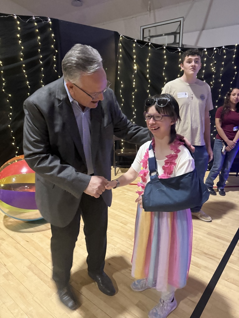 Tim Bryant, a school board member in a dark suit shakes hands with a smiling student wearing glasses, a colorful dress, and a pink lei, with one arm in a sling. Other students and a backdrop with string lights and tropical decorations are visible in the background.