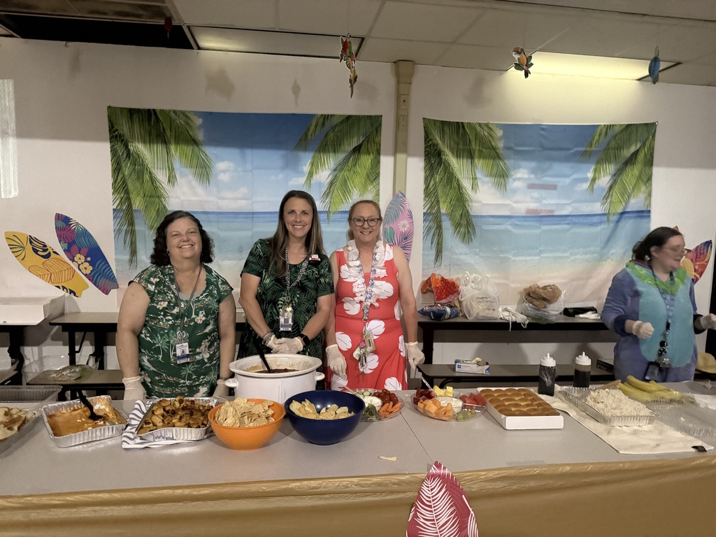 Richbourg teachers stand behind a table serving food during a school prom, with a beach‑themed backdrop in a school gymnasium.
