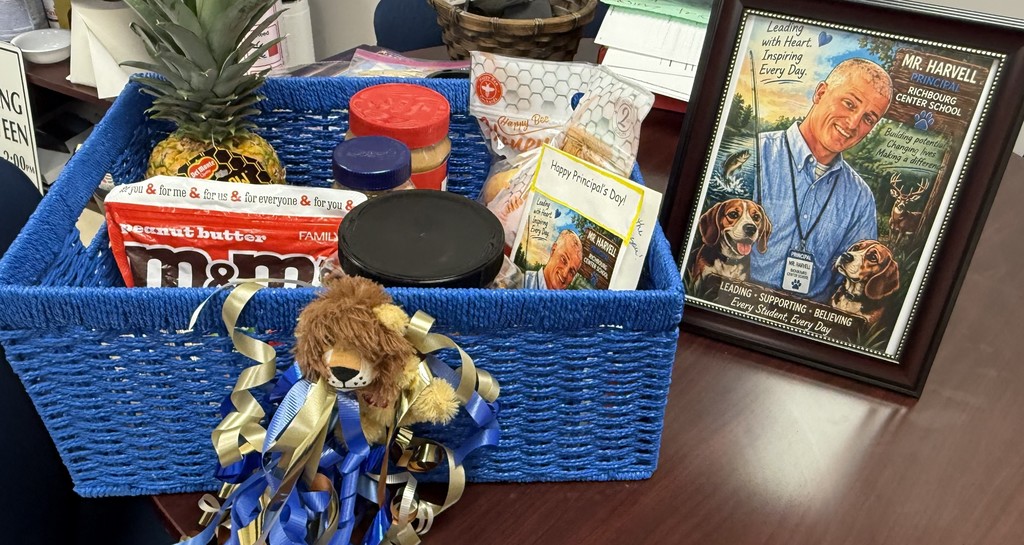 A blue gift basket filled with snacks, including peanut butter, candy, and a pineapple, sits on a desk. Next to it is a framed illustration of the school principa, Mr. Harvelll with outdoor scenes and two dogs, along with a card celebrating Principal  Day.