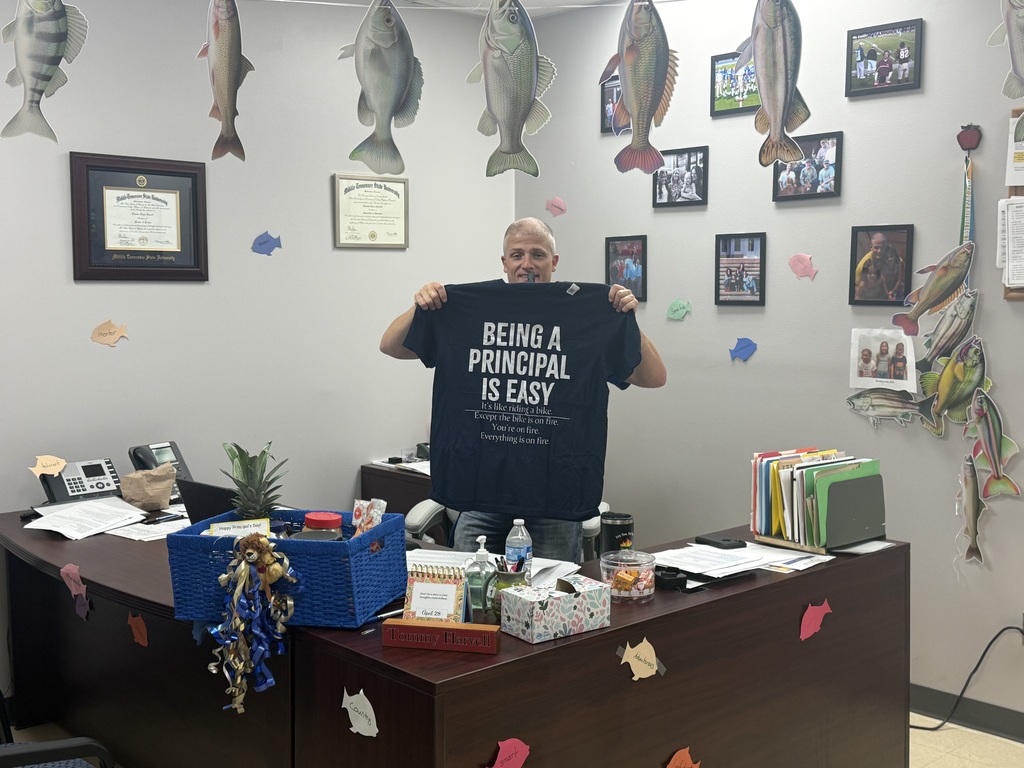 Richbourg principal, Mr. Harvell  stands behind his desk in a decorated office, holding up a dark-colored T-shirt with a humorous message about being a principal. The office is decorated with fish cutouts hanging from the ceiling and framed photos on the wall, reflecting a fishing theme.